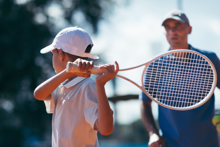 Trainer und Schüler Ein Junge hält einen Tennisschläger, während ein Trainer im Hintergrund zusieht.